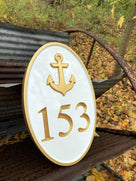 Side view of oval house number sign painted white and gold.  an anchor is the emblem over the number.